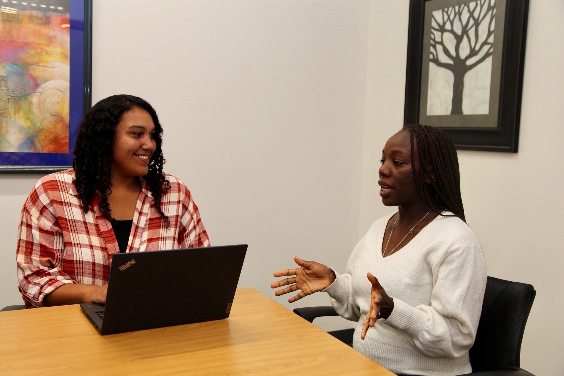 Two women talk at an office table with a laptop.